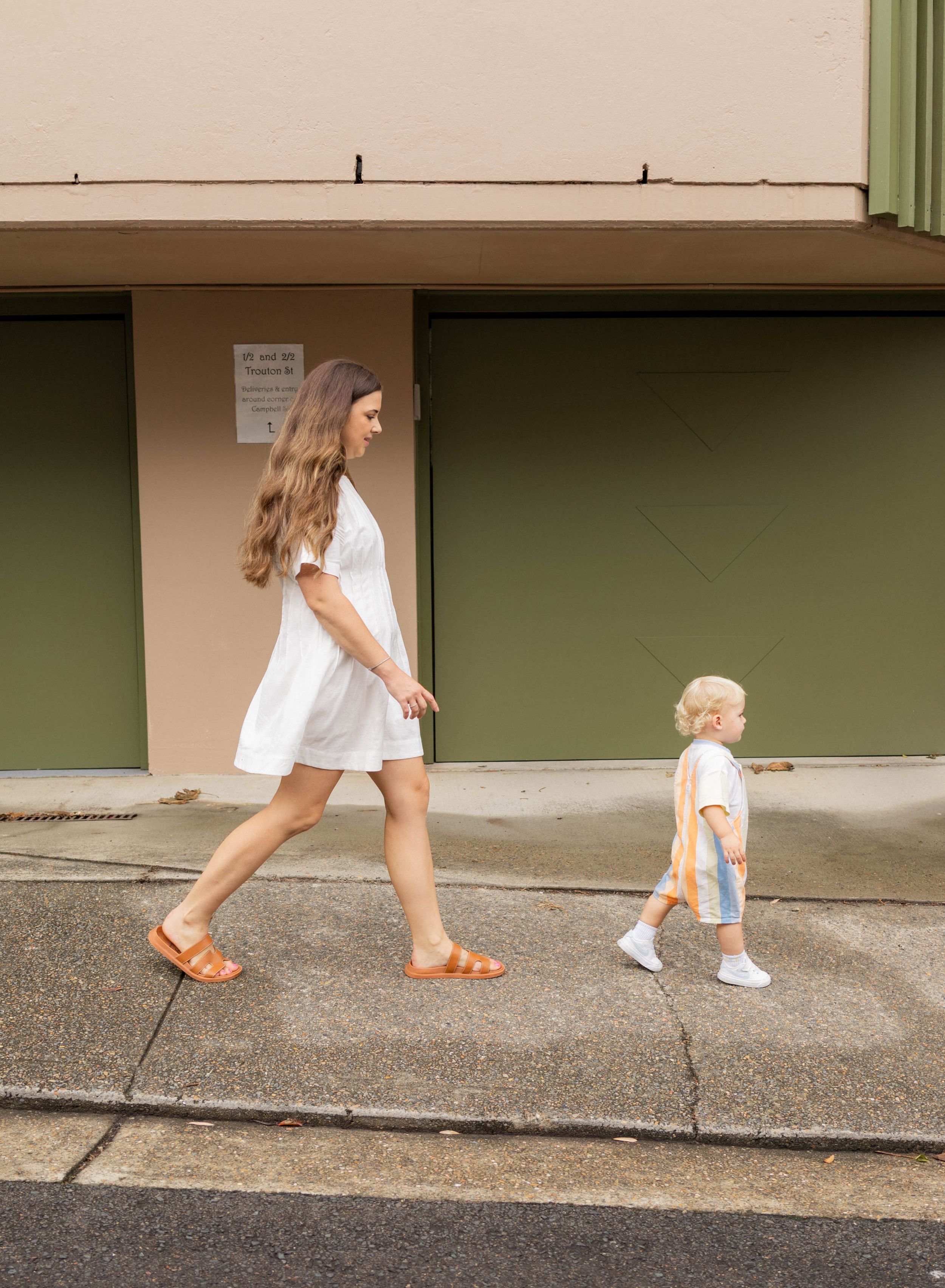 Young mother in white dress walking with toddler child on urban sidewalk.