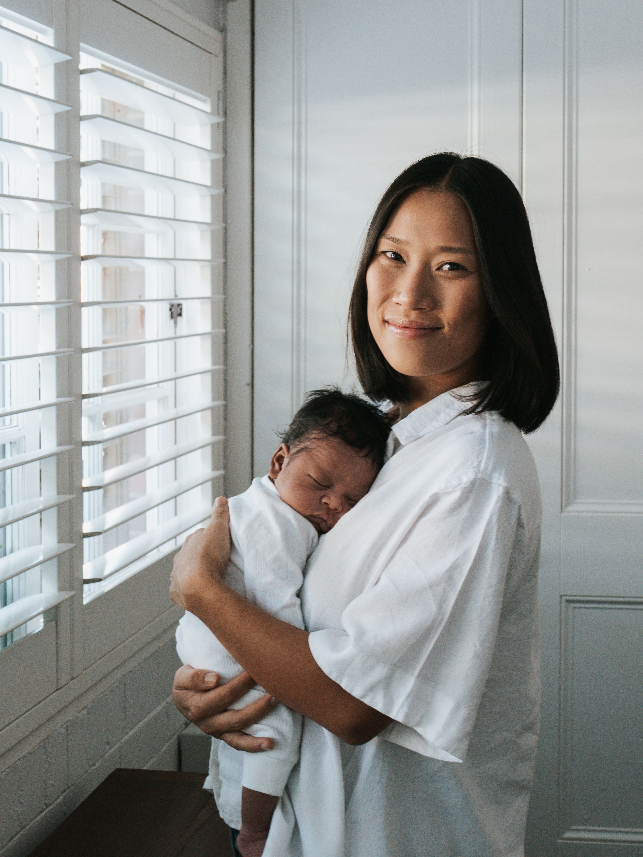 Asian mother in white shirt holding sleeping newborn baby by window indoors.