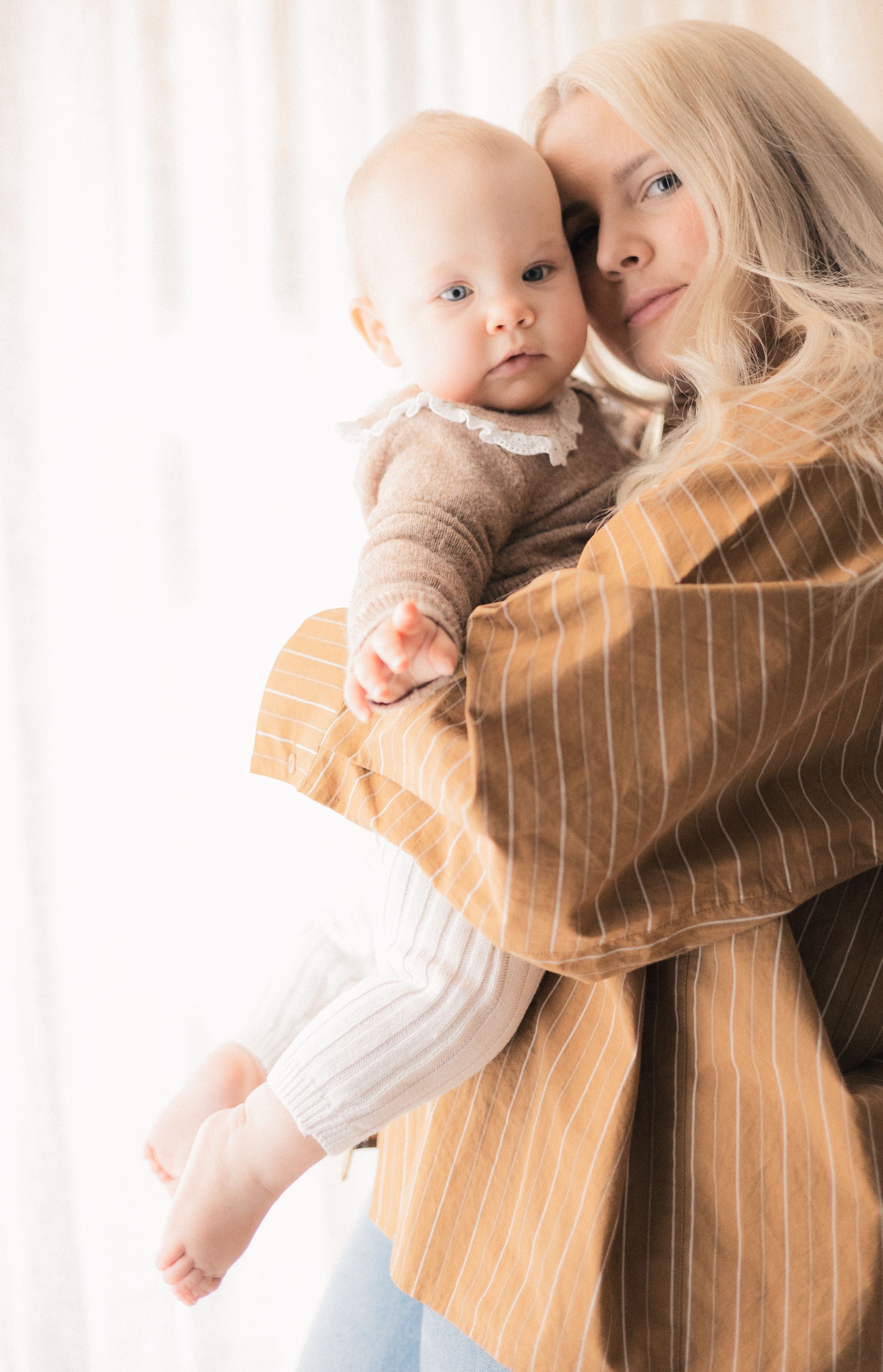 Blonde woman in striped camel blazer tenderly holds a baby in brown outfit near sheer white curtains.
