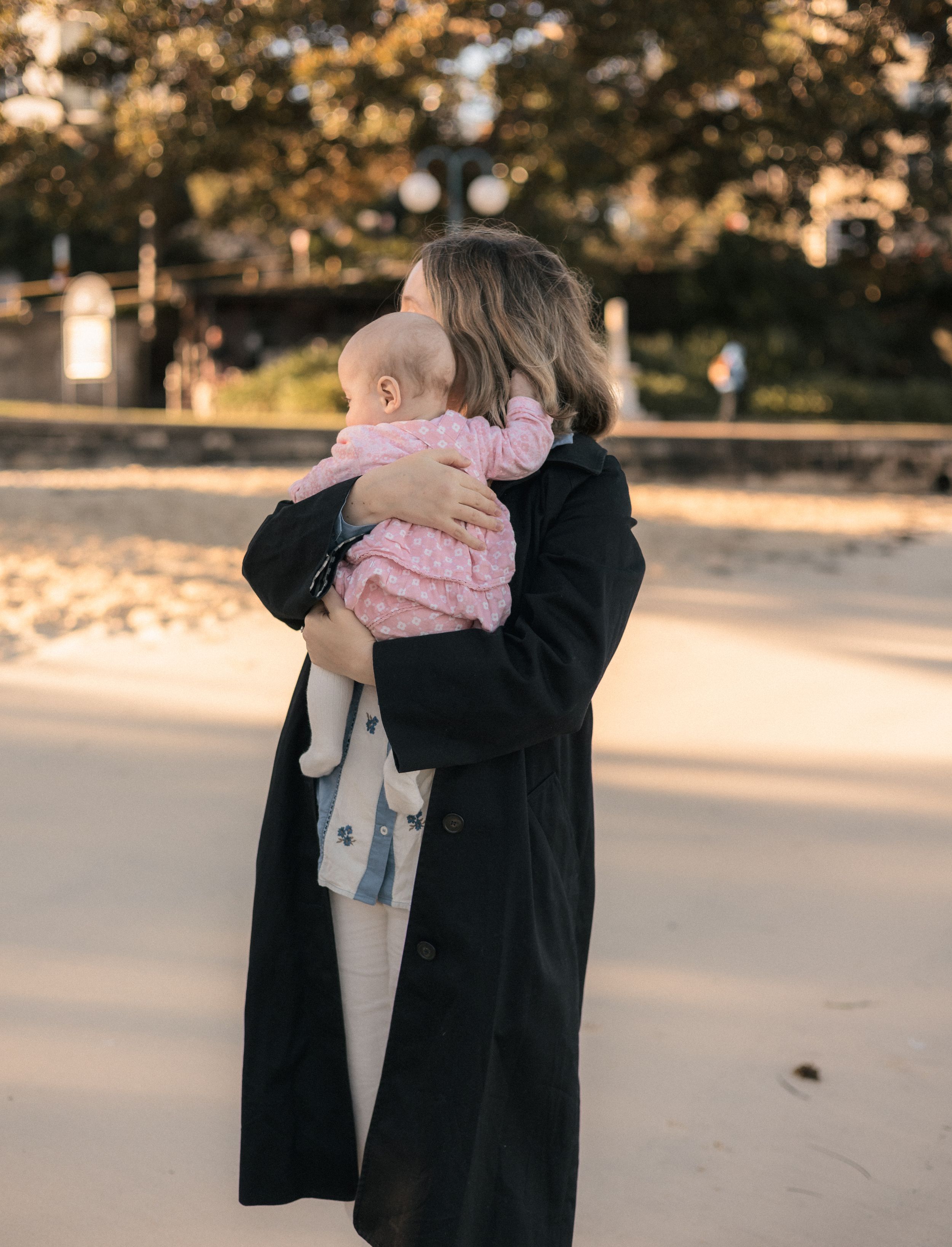 Mother in black coat cradling infant baby on sandy beach path at golden hour.