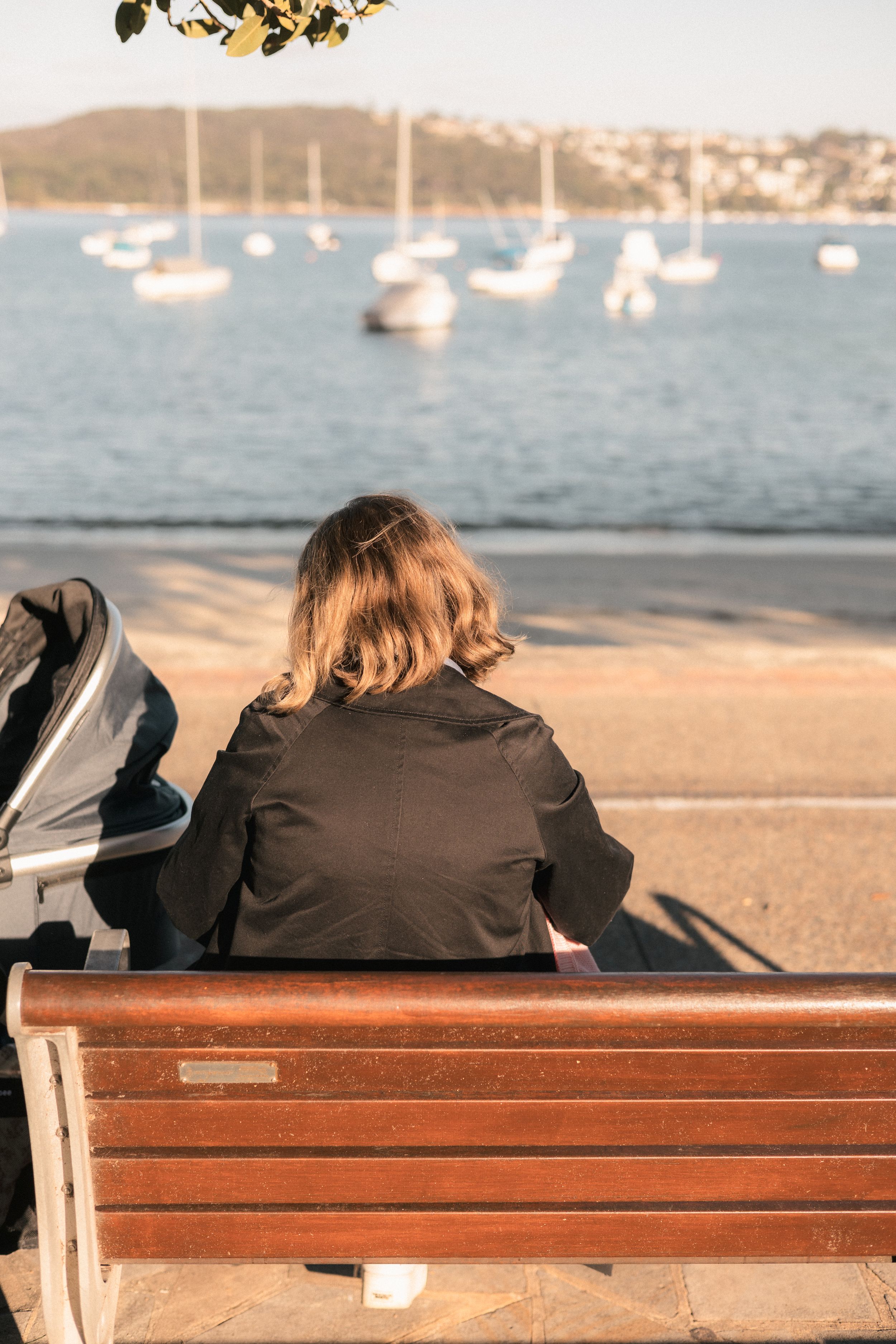 Woman in black jacket sitting on wooden bench overlooking harbor with sailboats and hills in background breastfeeding her baby