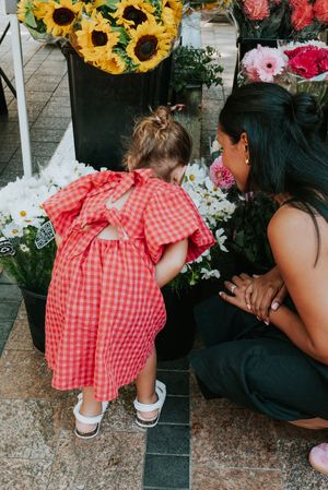 A toddler in a red gingham dress smells white flowers at a market stall with a woman crouching beside her.
