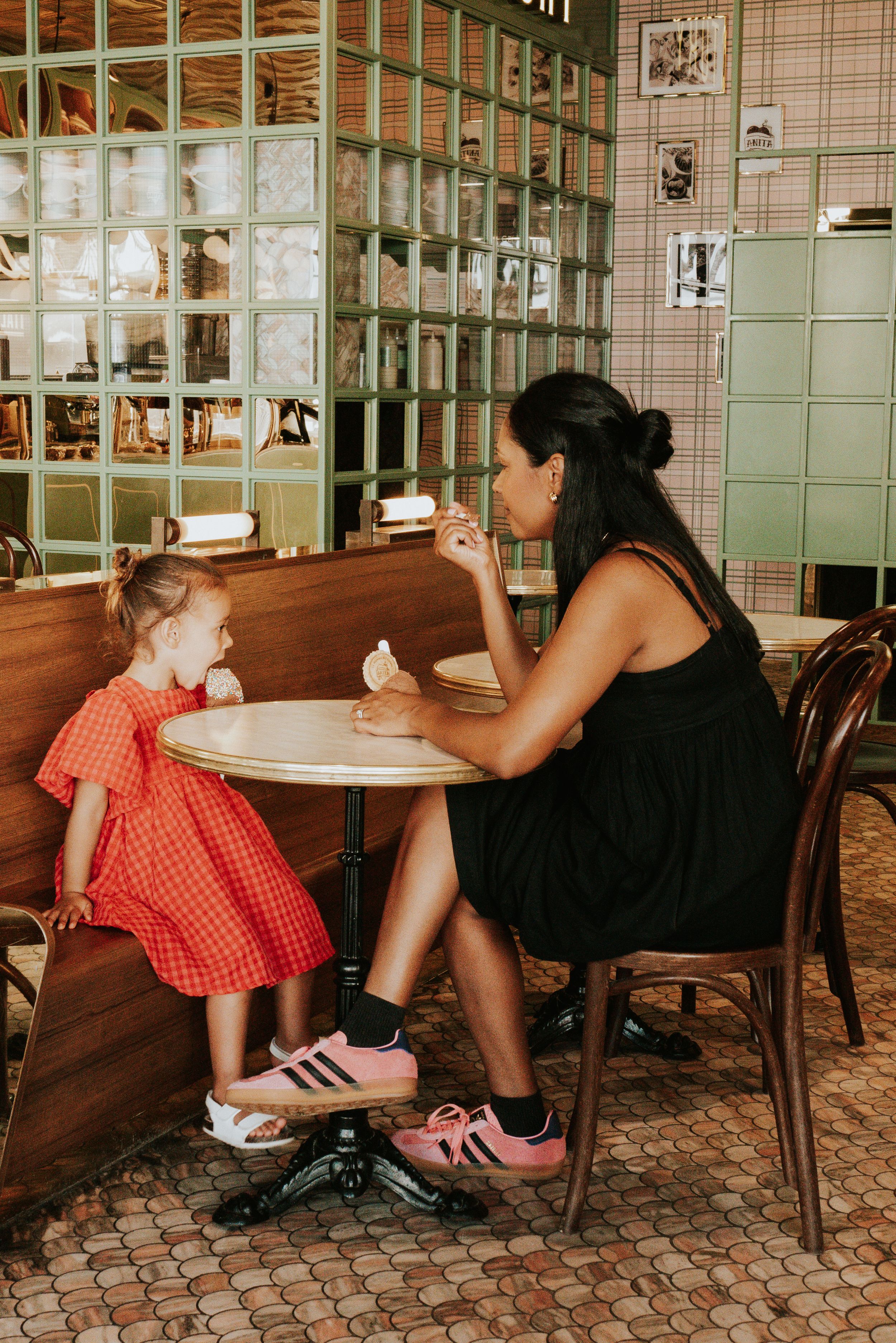 Woman in black dress shares ice cream with young girl in red dress at a cafe table.