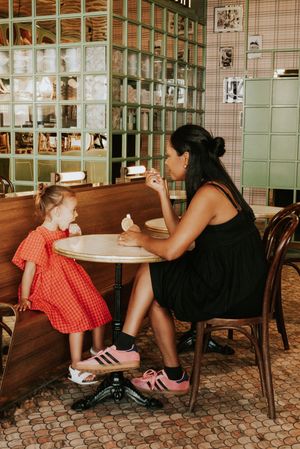Woman in black dress shares ice cream with young girl in red dress at a cafe table.
