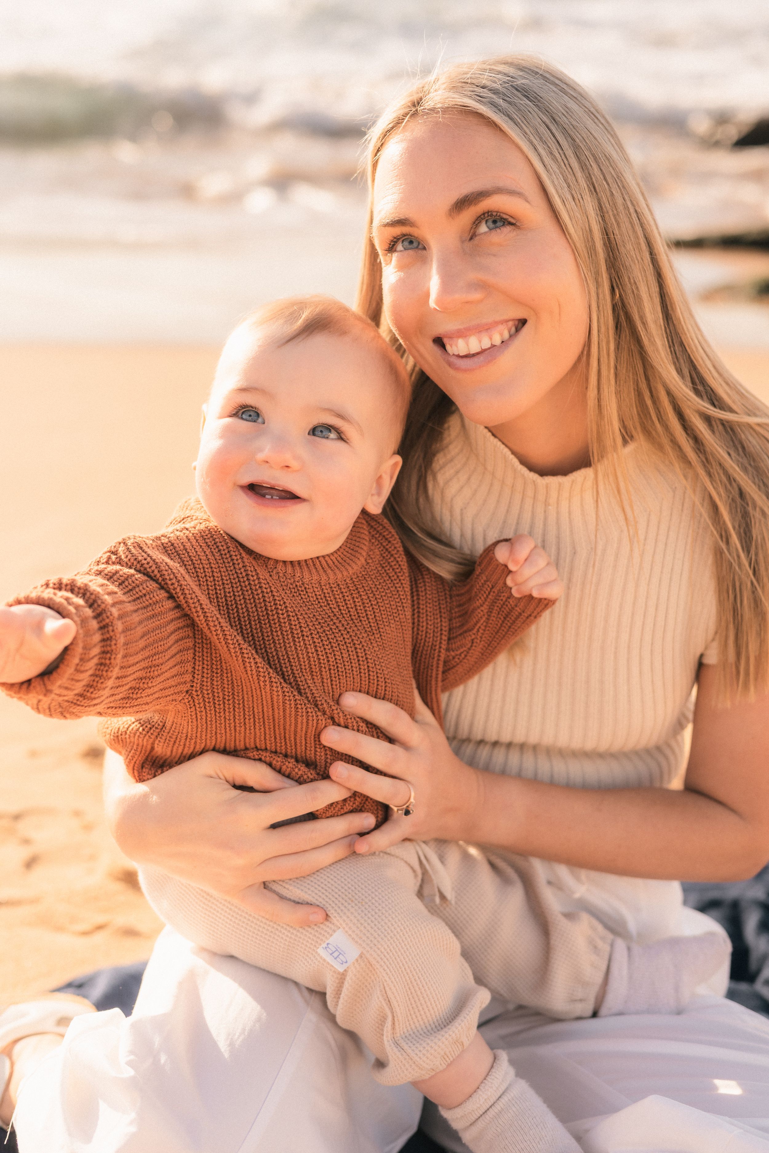 Smiling blonde mother holding happy baby on sunny beach, both wearing warm knit sweaters.