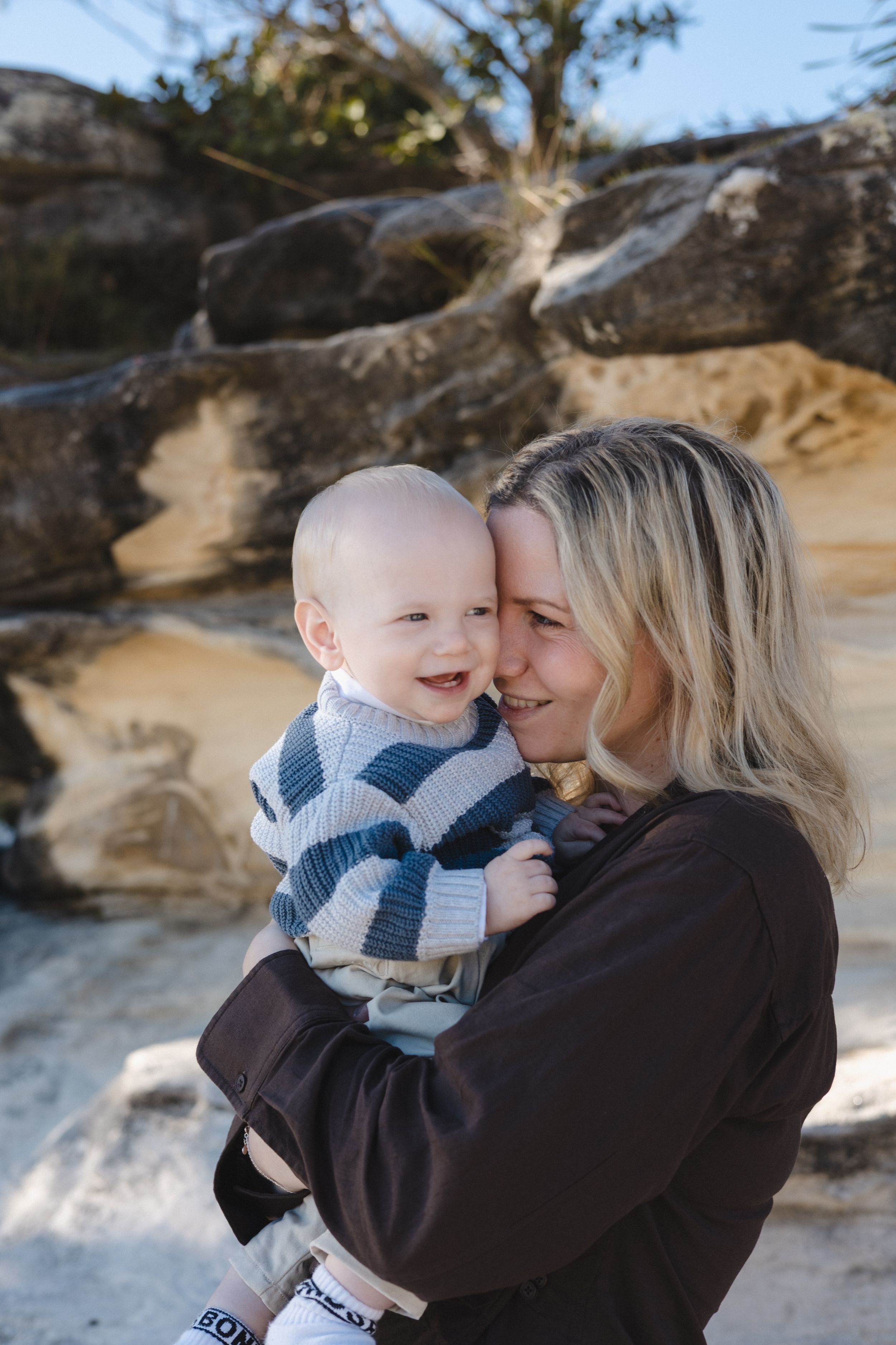 A smiling blonde woman in a brown top holds a happy baby in a striped sweater against rocky background.