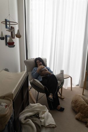 A woman sits in a rocking chair cradling a newborn in a softly lit nursery with a mobile and stuffed animals.