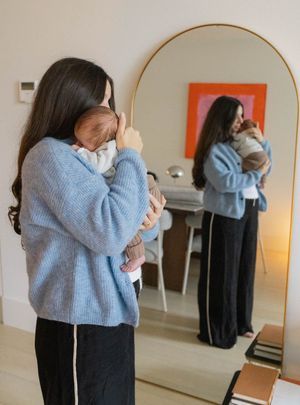 Woman in blue sweater cradling newborn baby, reflected in large arched gold mirror in modern home.
