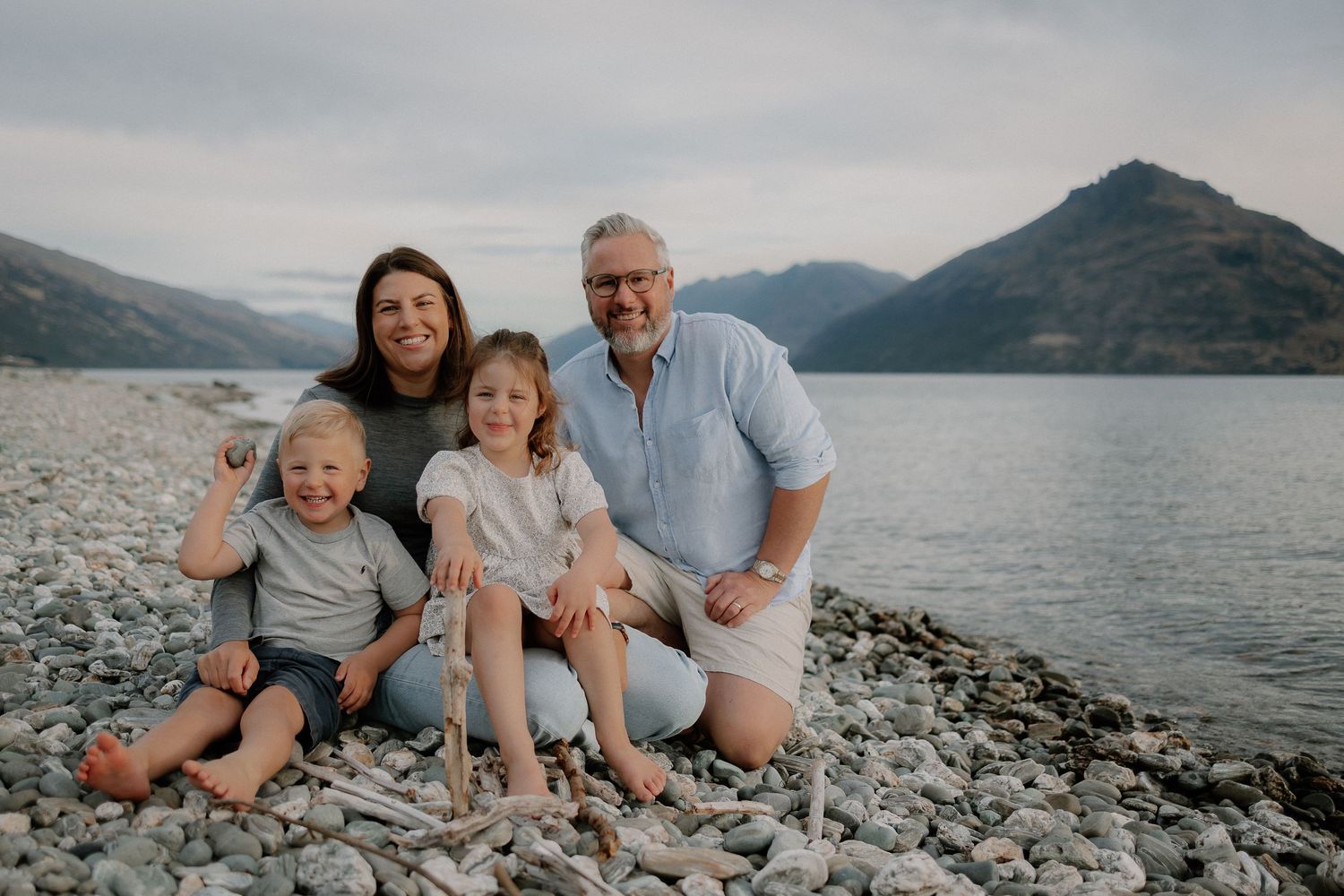 Family of four smiling on a rocky beach with a scenic lake and mountain backdrop.