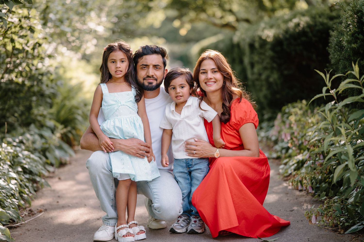 Happy family of four posing on a garden path surrounded by lush greenery, parents with two young children smiling.