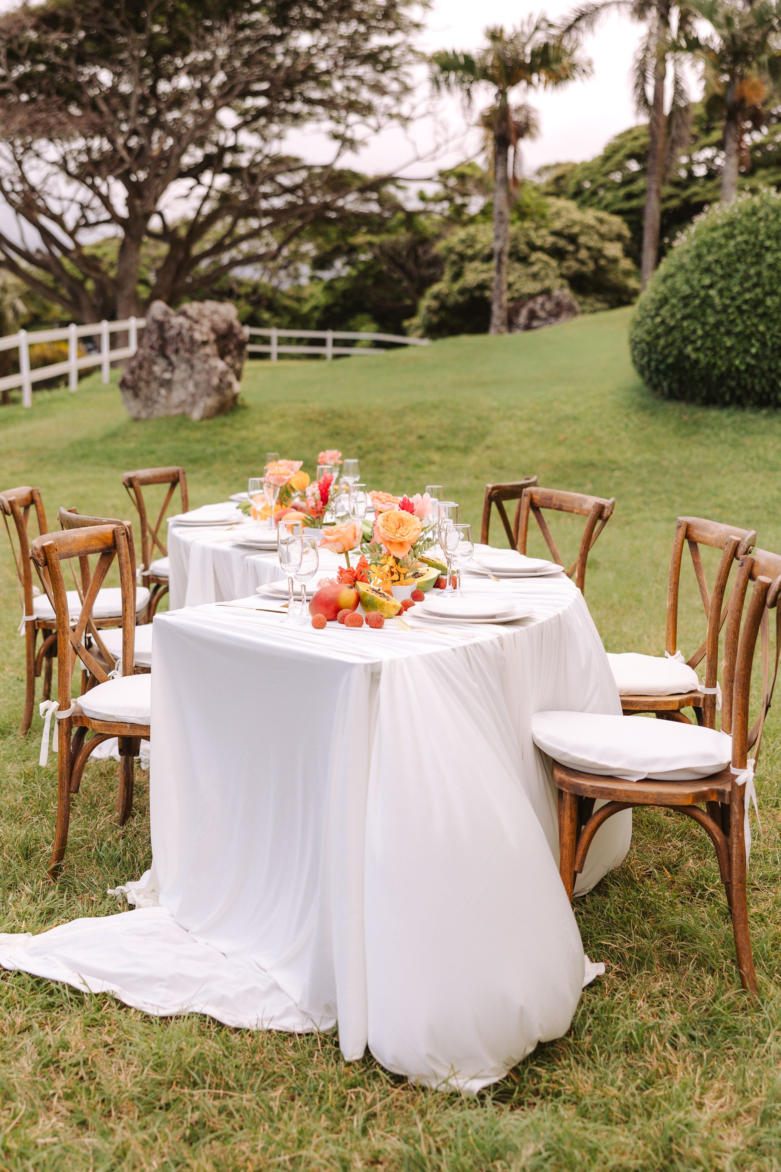 Elegant outdoor wedding reception table with white linen, floral centerpiece, and wooden cross-back chairs on lush green lawn.