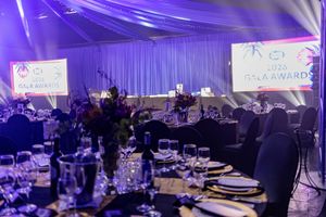 Elegant gala awards dinner setup with purple lighting, floral centerpieces, and branded screens in a draped venue.