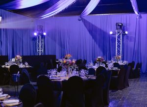 Elegant banquet hall with blue lighting, round tables with floral centerpieces, black chair covers, and draped ceiling decor.