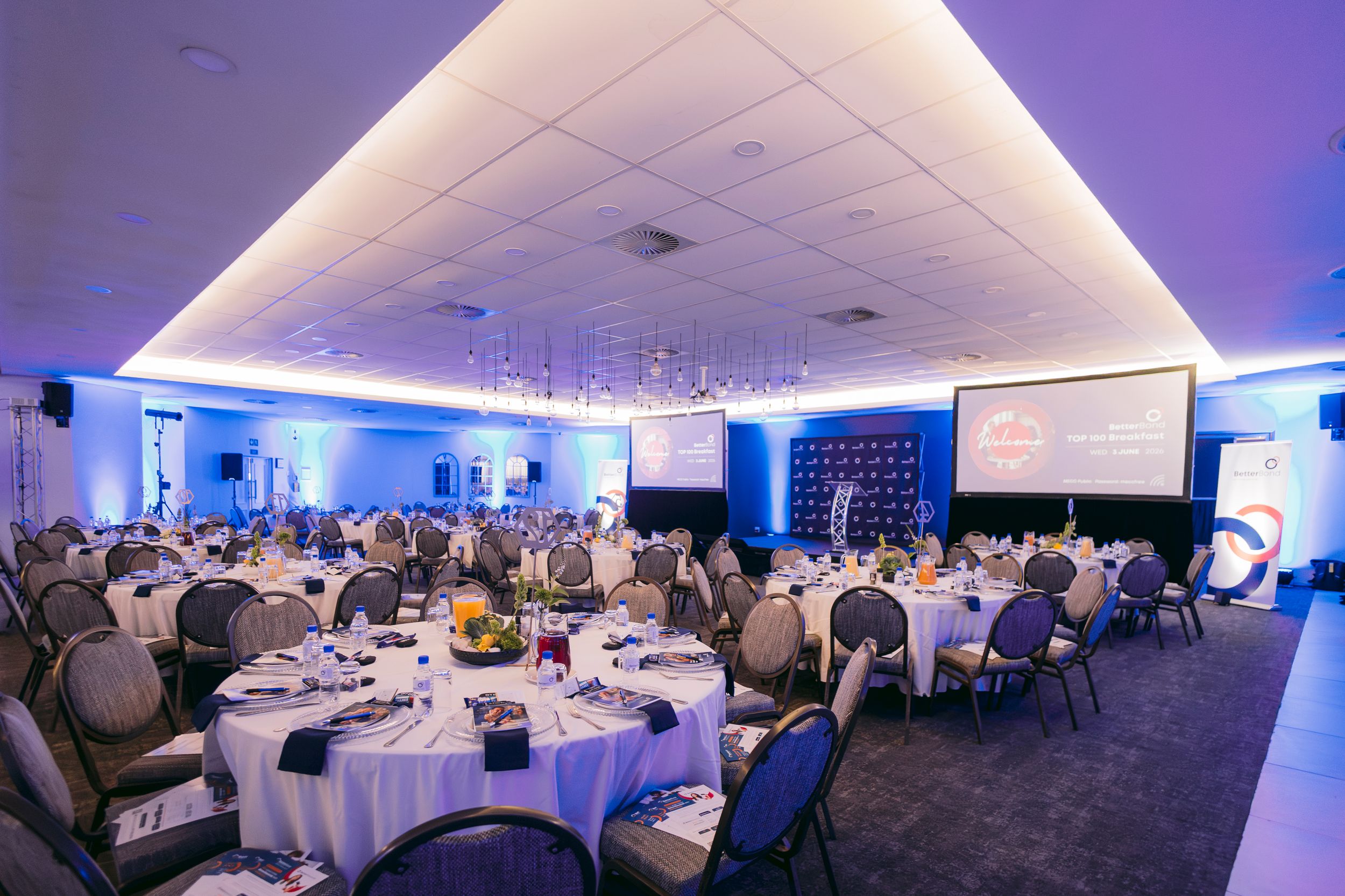 Elegant banquet hall with round tables, white linens, blue ambient lighting, and a projection screen for corporate events.