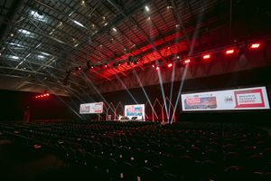Large empty conference hall with red stage lighting, presentation screens, and rows of red seats in a modern venue.