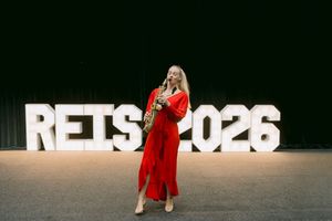 Woman in red dress playing saxophone in front of illuminated RETS 2026 sign on dark stage.