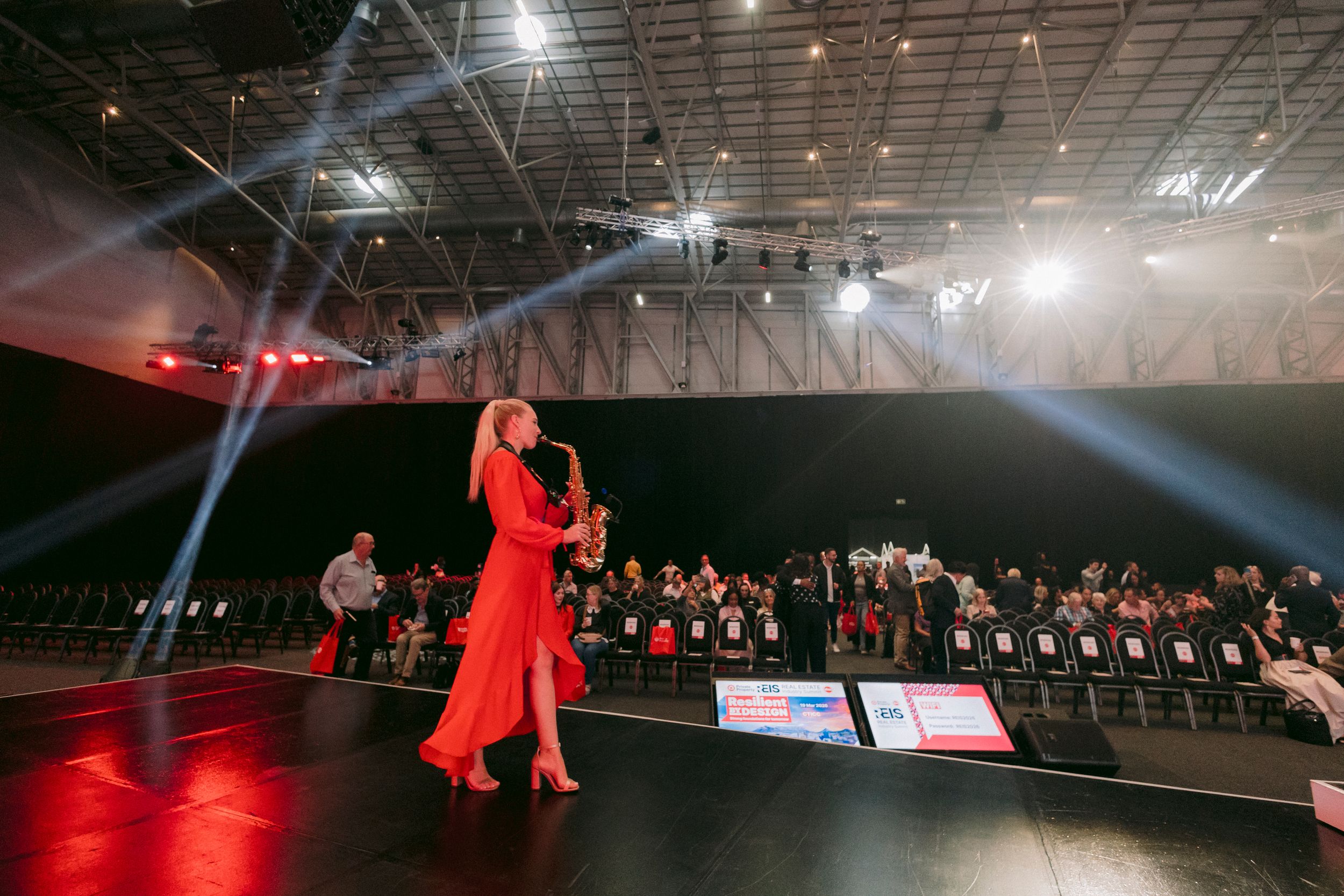Woman in red dress plays saxophone on stage with dramatic lighting at a large indoor event venue.