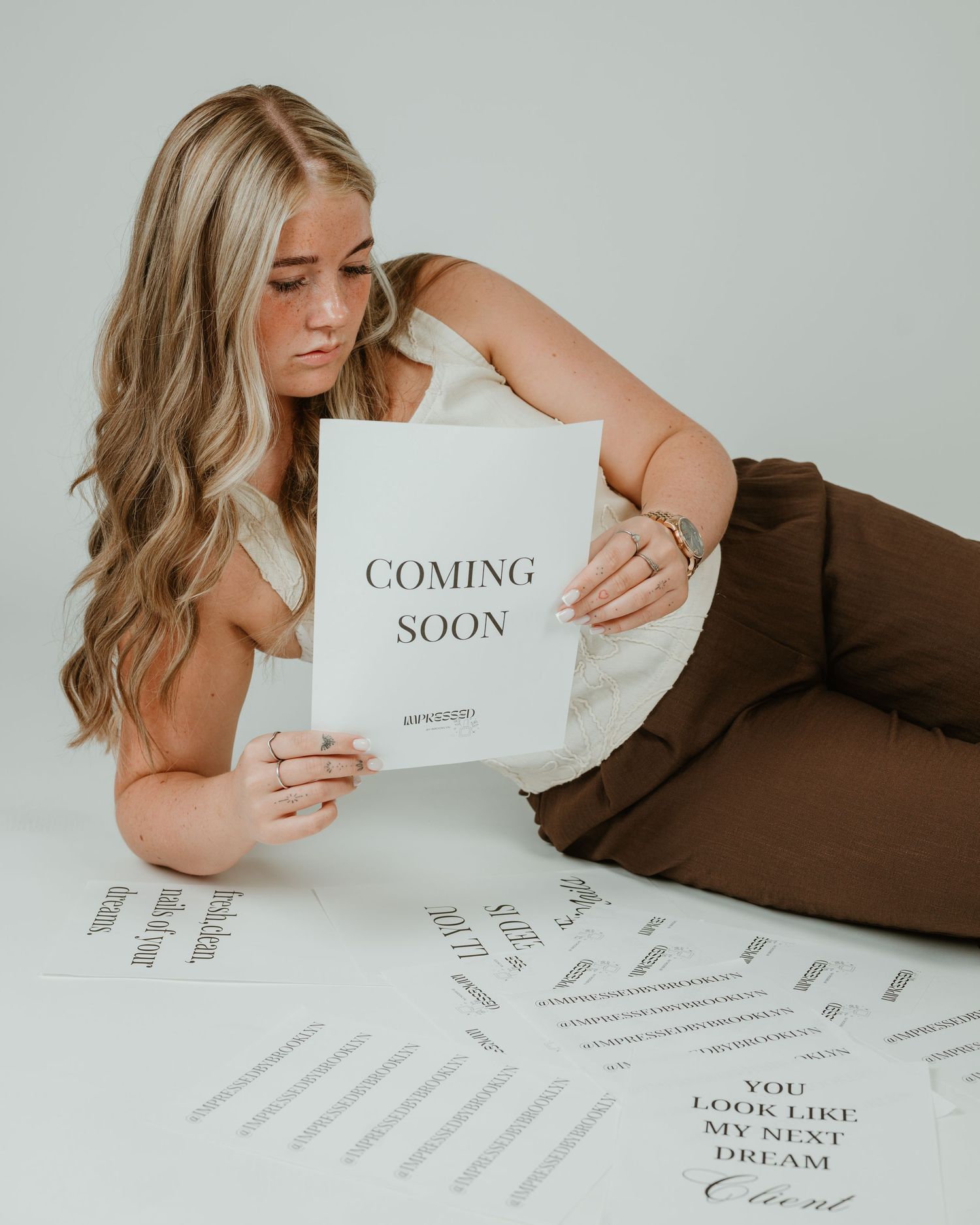 Young blonde woman in brown pants holds a Coming Soon sign while lounging on floor surrounded by printed cards.