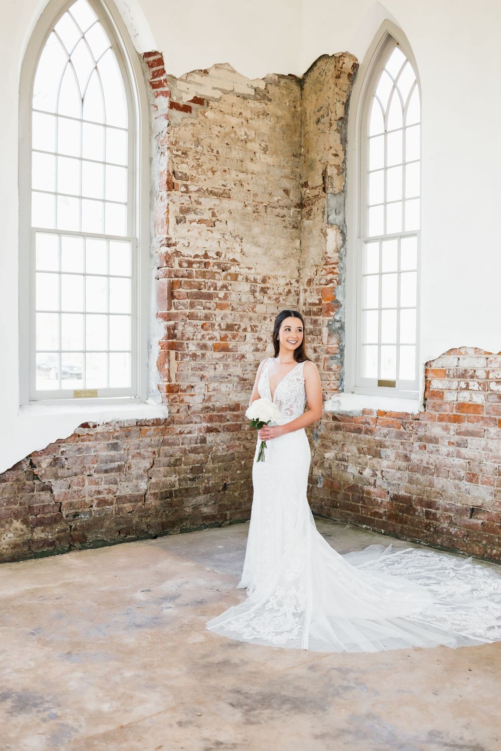 Bride in lace wedding gown with train poses inside rustic brick church with arched windows.