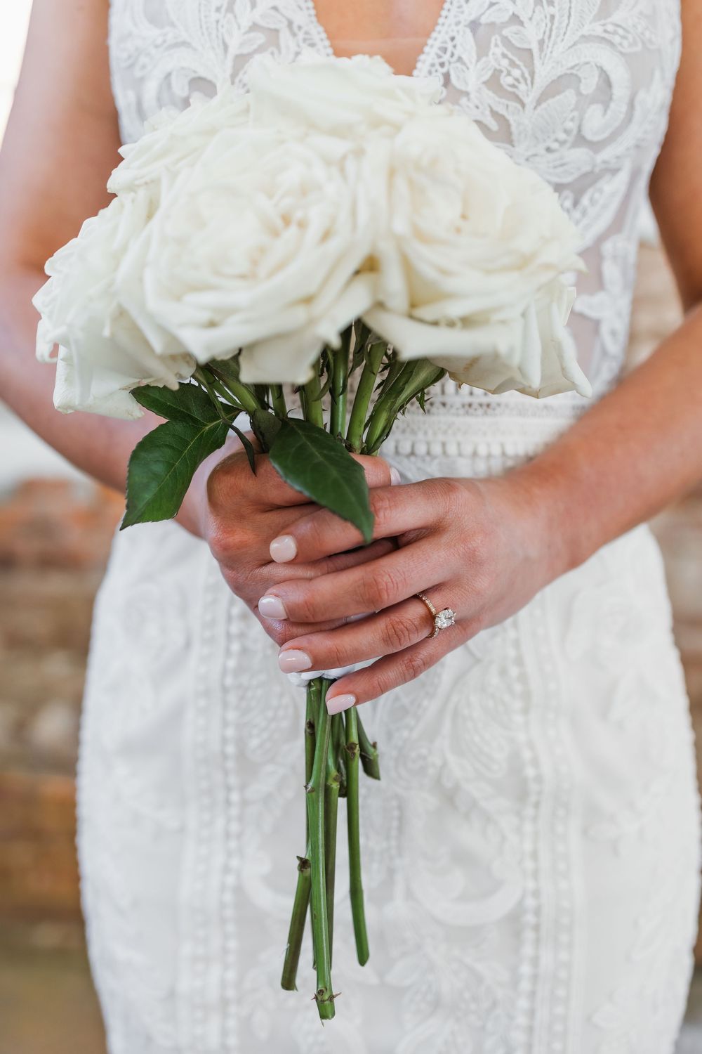 Bride in lace wedding dress holding elegant white rose bouquet with green stems, featuring diamond engagement ring.