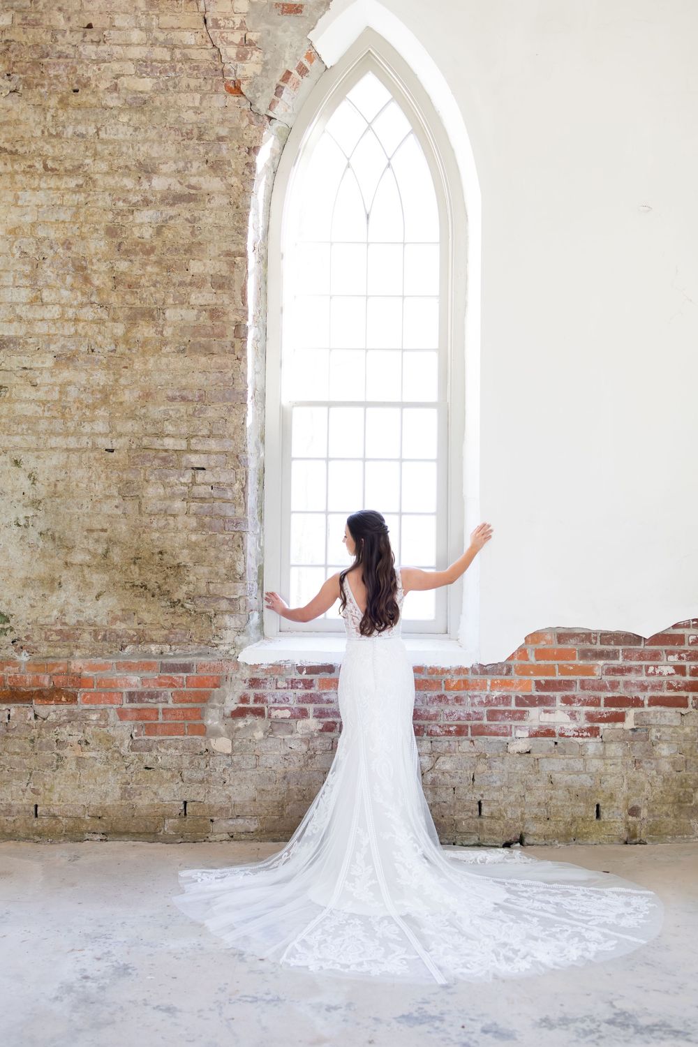 Bride in white lace gown with long train stands before Gothic arch window in rustic brick chapel.