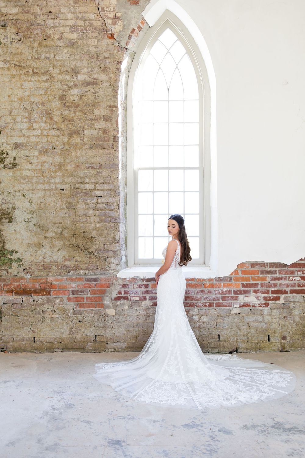 Bride in white lace mermaid gown with train poses by arched window in rustic brick chapel.