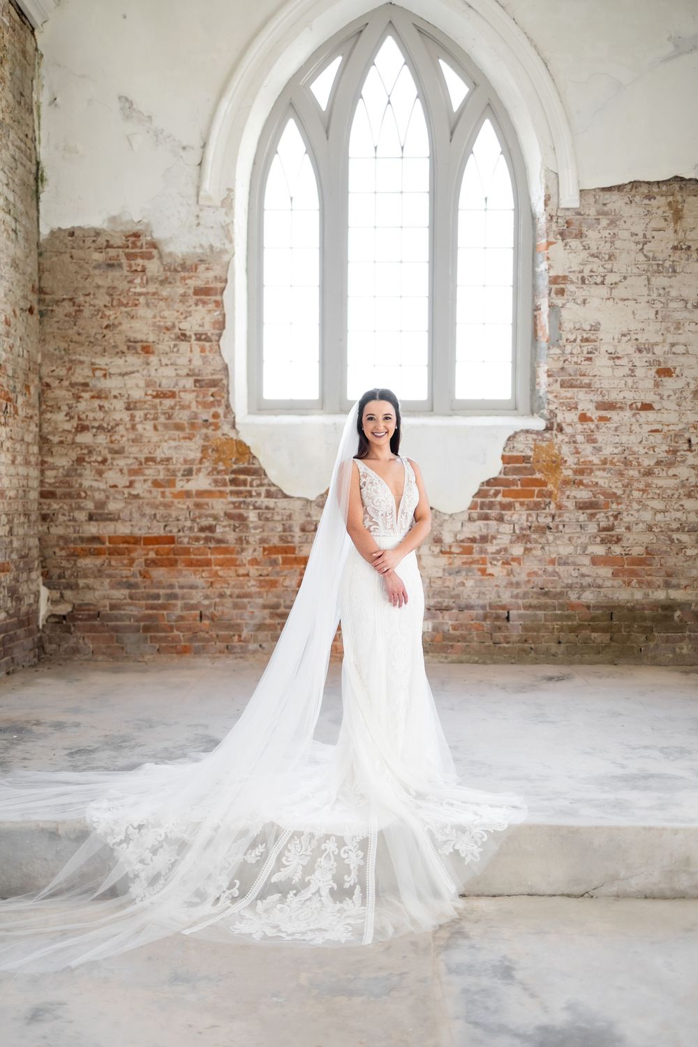 Bride in white lace gown with flowing veil stands before gothic arched window in rustic brick venue.