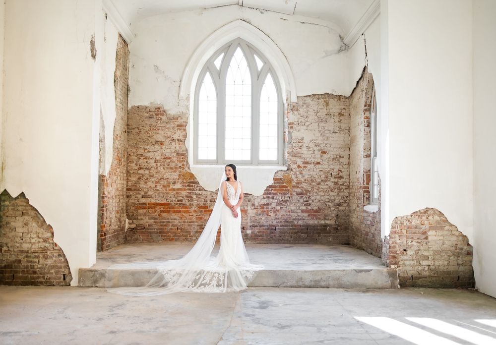 Bride in white gown poses in rustic abandoned chapel with exposed brick walls and arched window.