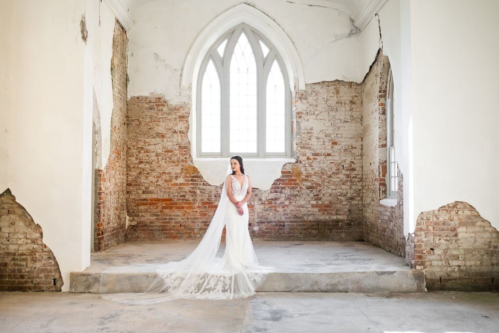 Bride in white gown with long veil poses in rustic brick chapel with arched window.