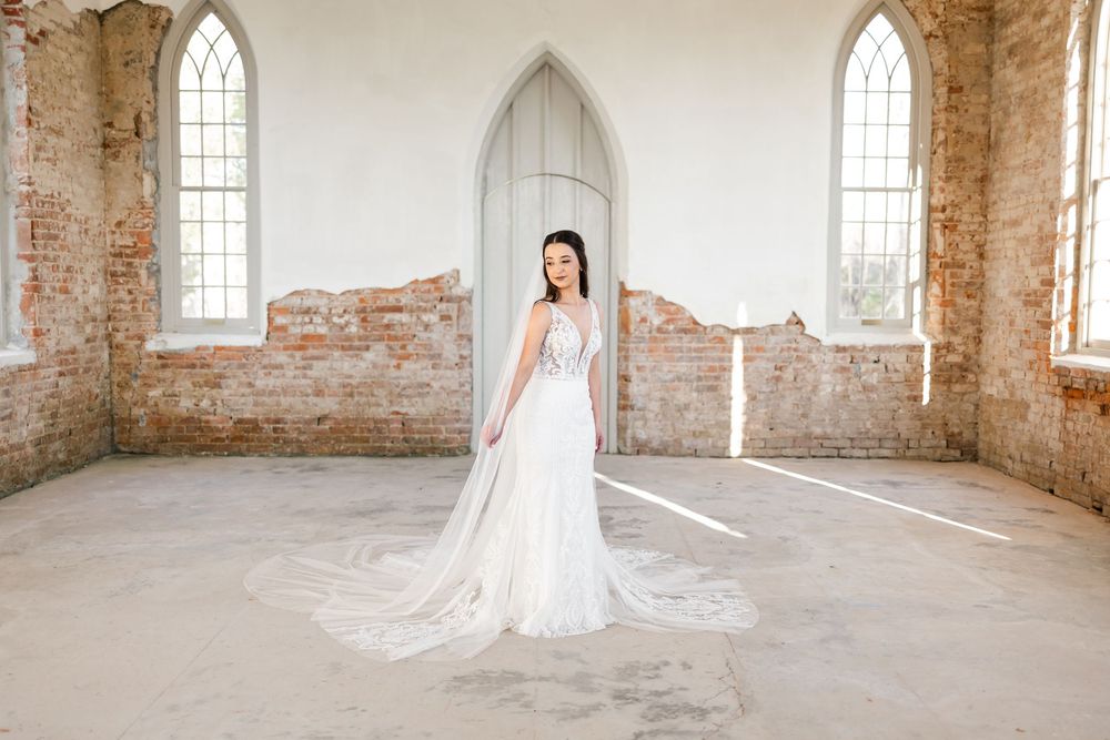 Bride in elegant white gown with long train stands in rustic brick chapel with arched windows and natural light.