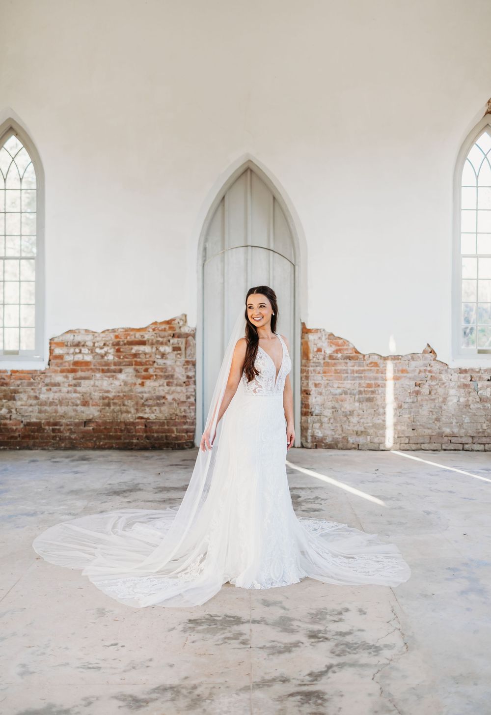Elegant bride in fitted white gown with cathedral veil poses in sunlit rustic brick chapel interior.