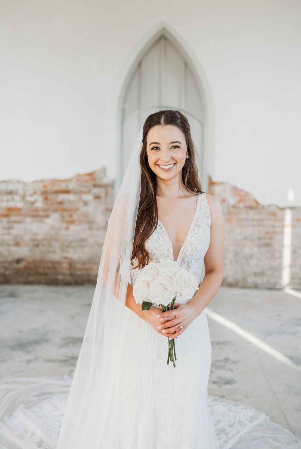 Smiling bride in V-neck wedding dress with veil holds white bouquet outside brick chapel.
