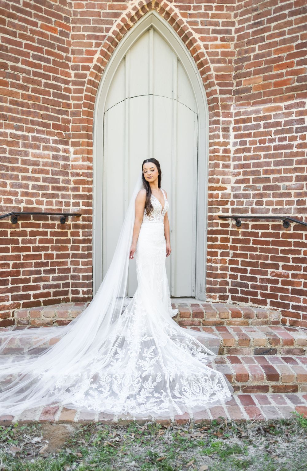 Bride in elegant white mermaid gown with lace train and veil stands before gothic arched door on brick church exterior.