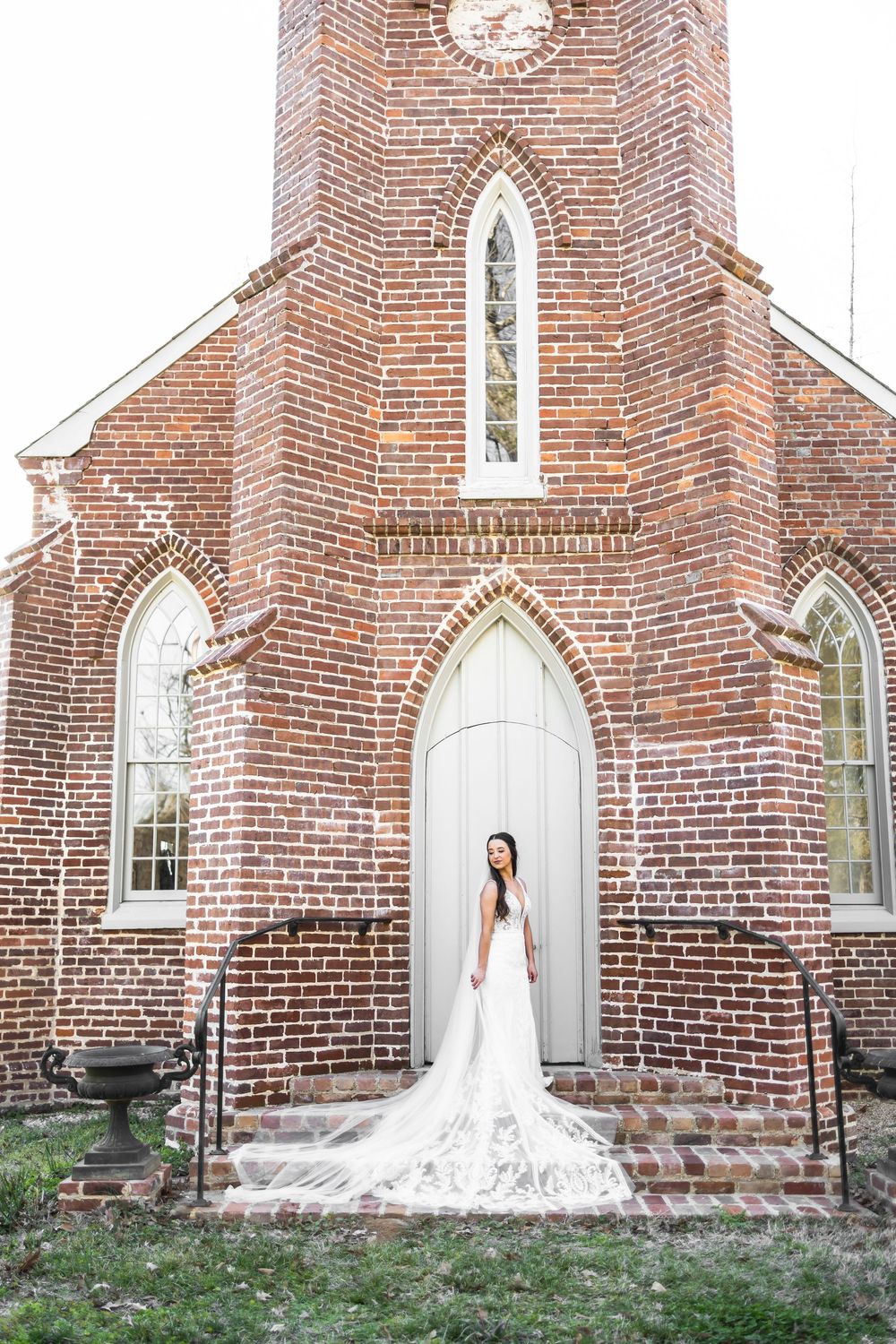 Bride in lace gown with cathedral train stands before ornate red brick church entrance.