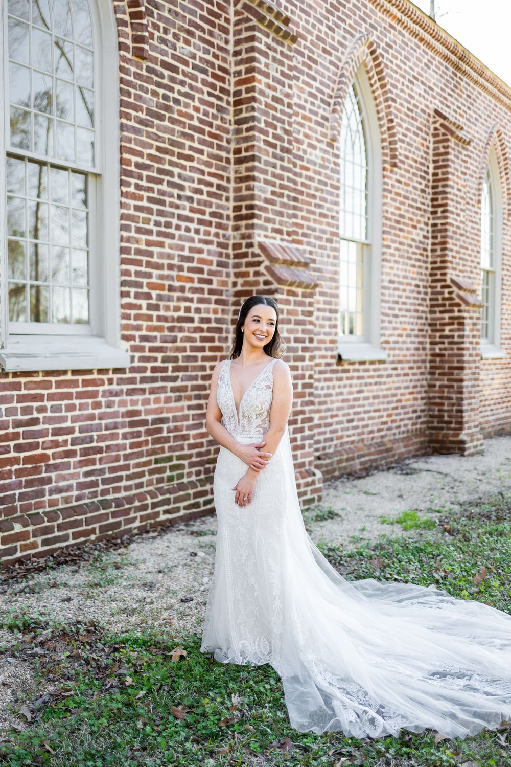 Elegant bride in fitted lace wedding dress with long train poses beside historic brick church.