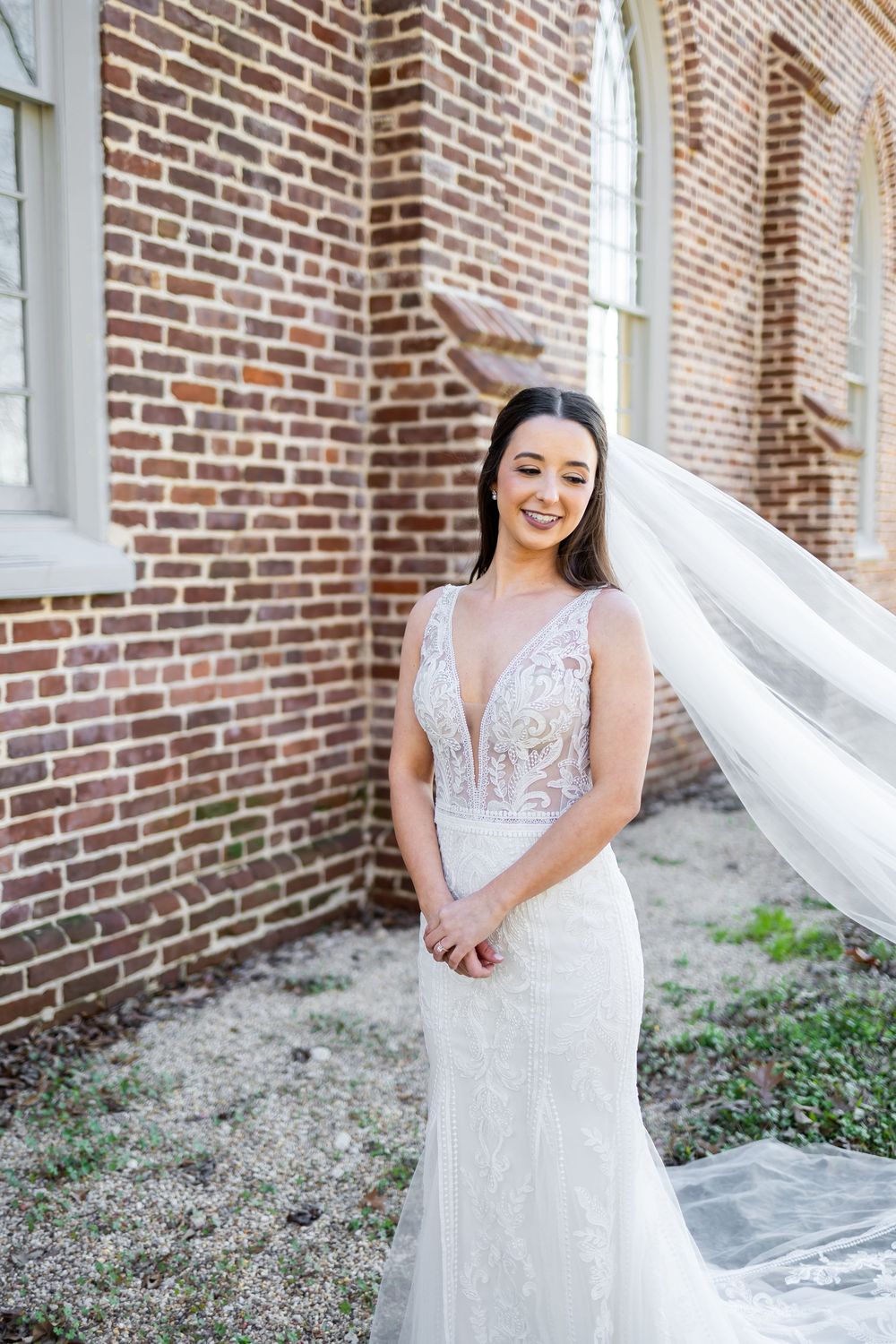 Smiling bride in lace plunging gown with flowing veil outdoors against historic brick building.