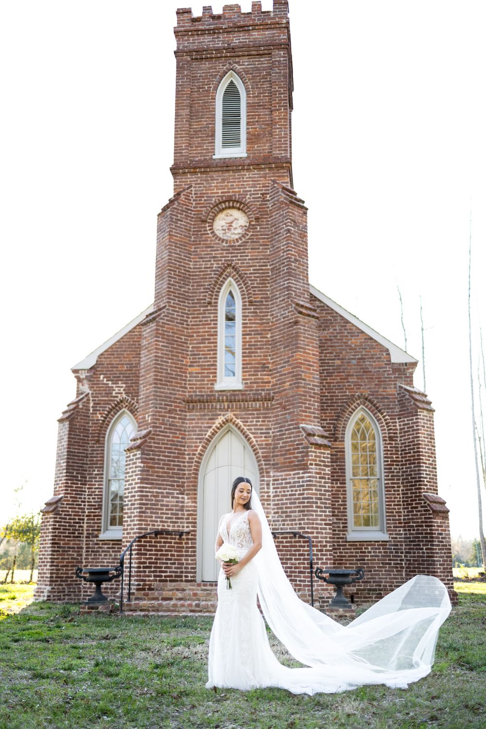 Bride in white mermaid gown with billowing veil stands before historic Gothic brick church exterior.