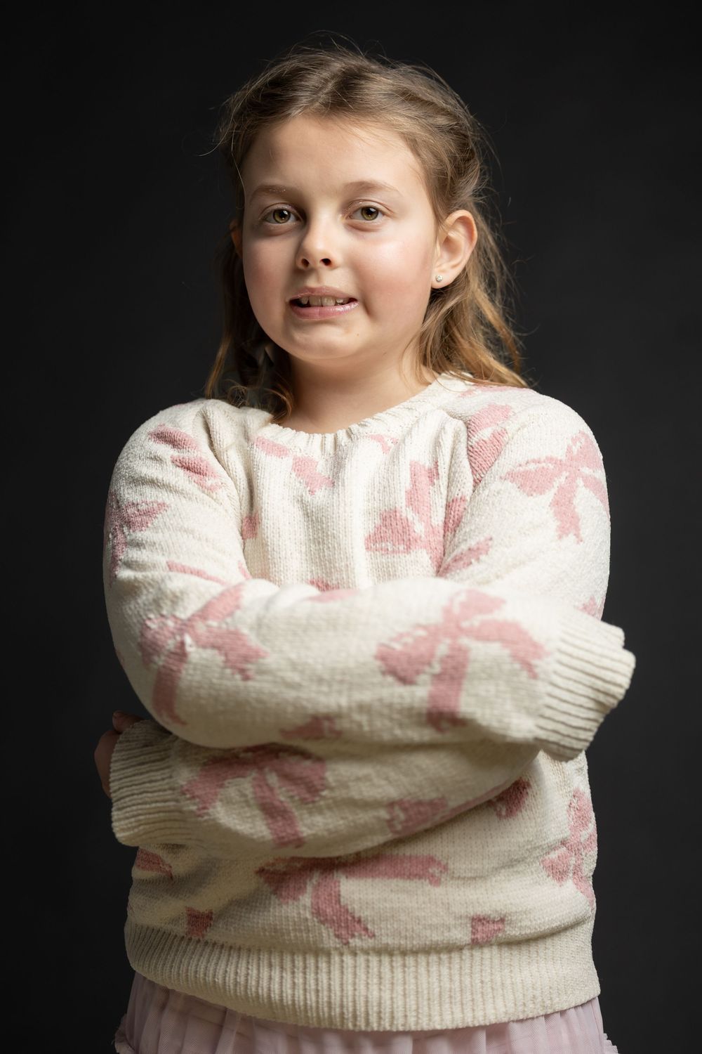 Young girl with arms crossed wearing a white and pink sweater posing against a dark background.