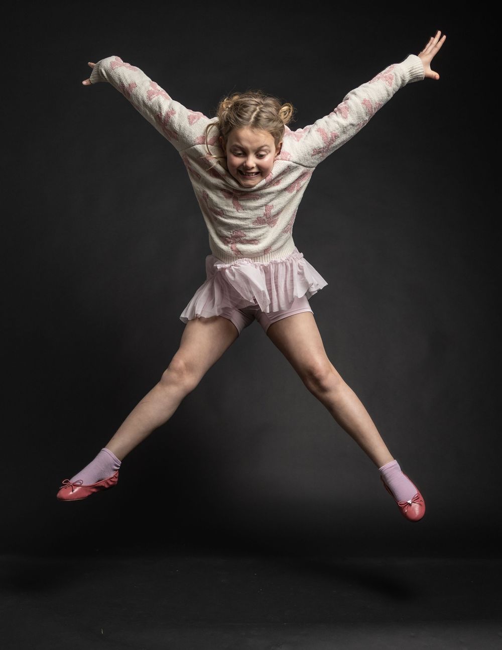 Energetic young girl jumping in star pose wearing pink tutu and floral sweater in studio.