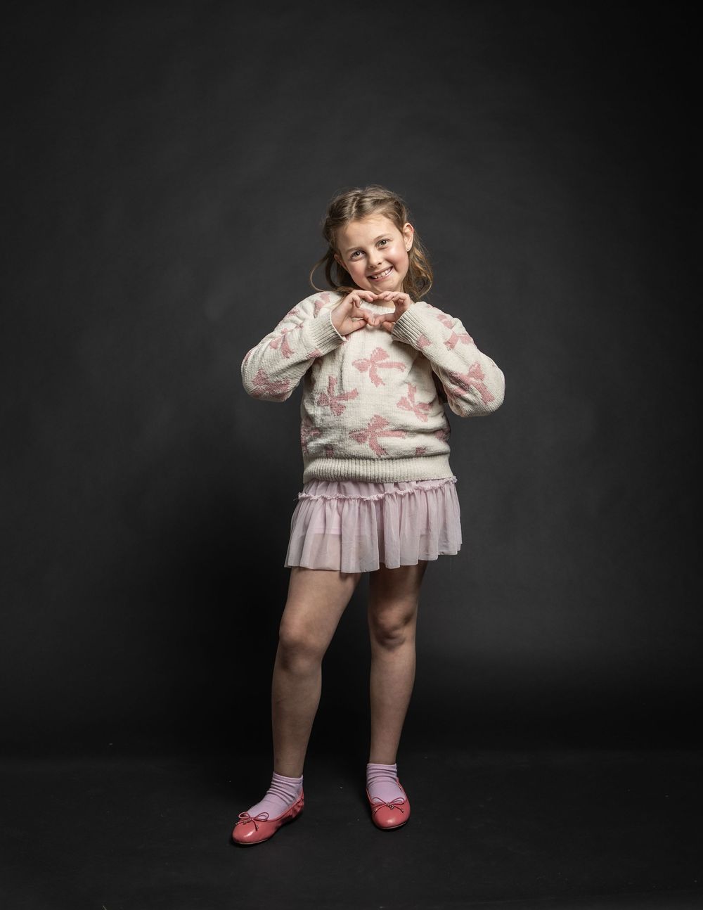 Adorable young girl making a heart shape with her hands, wearing floral sweater and pink skirt on dark background.