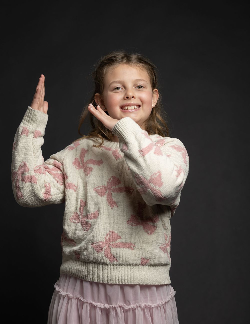 A smiling young girl in a pink bow-print sweater poses playfully with hands raised against a dark studio background.