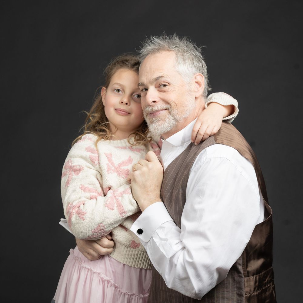 A young girl and an elderly man share a warm embrace, smiling together in a studio portrait against a dark background.
