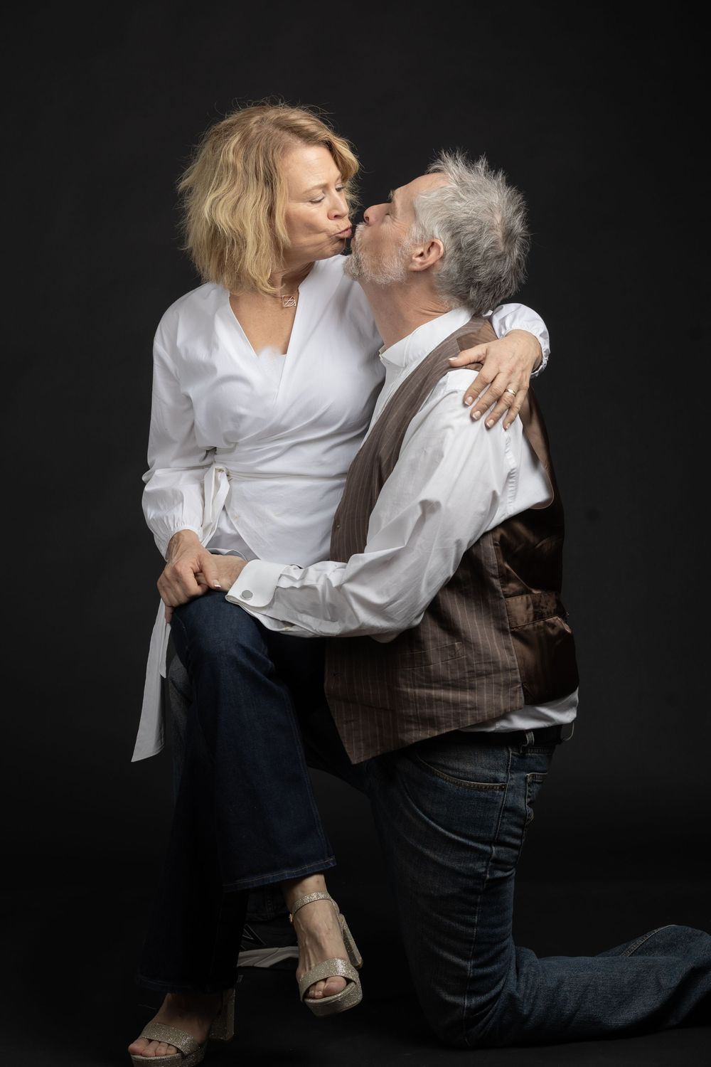 Mature couple sharing a romantic kiss, man kneeling in jeans and vest, woman in white blouse and jeans, dark studio background.