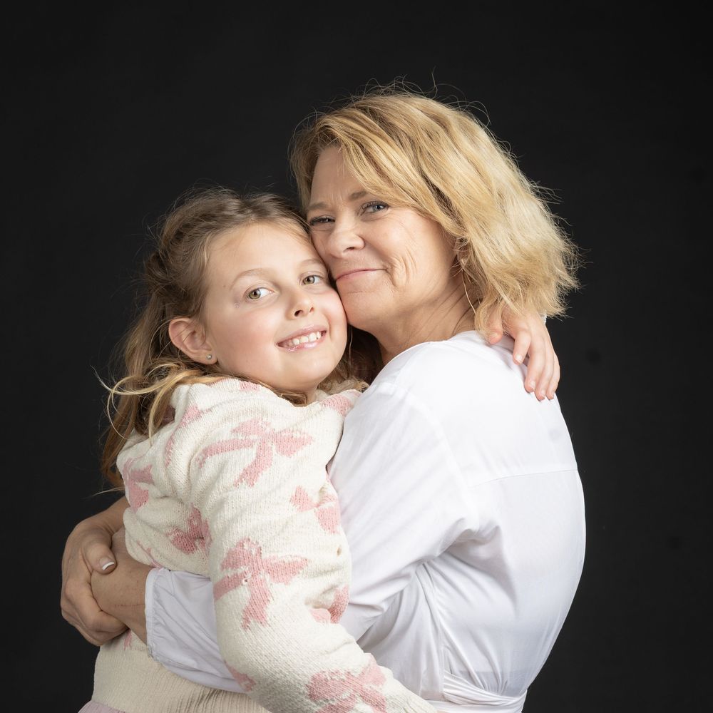 Blonde woman and young girl in warm embrace smiling together against dark studio background.