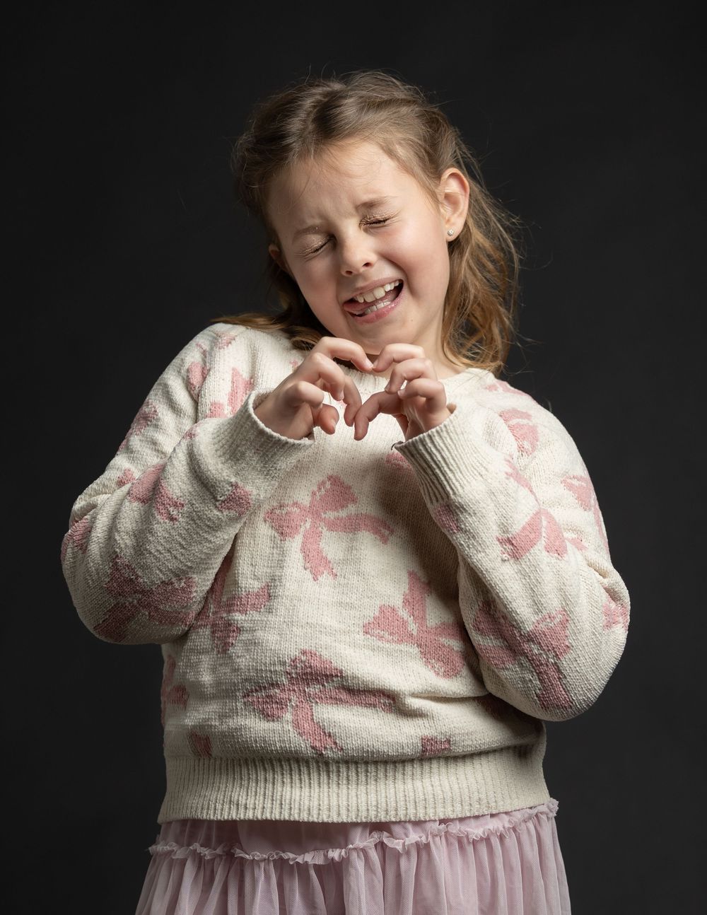 A young girl in a floral sweater makes a heart shape with her fingers, smiling with eyes closed.