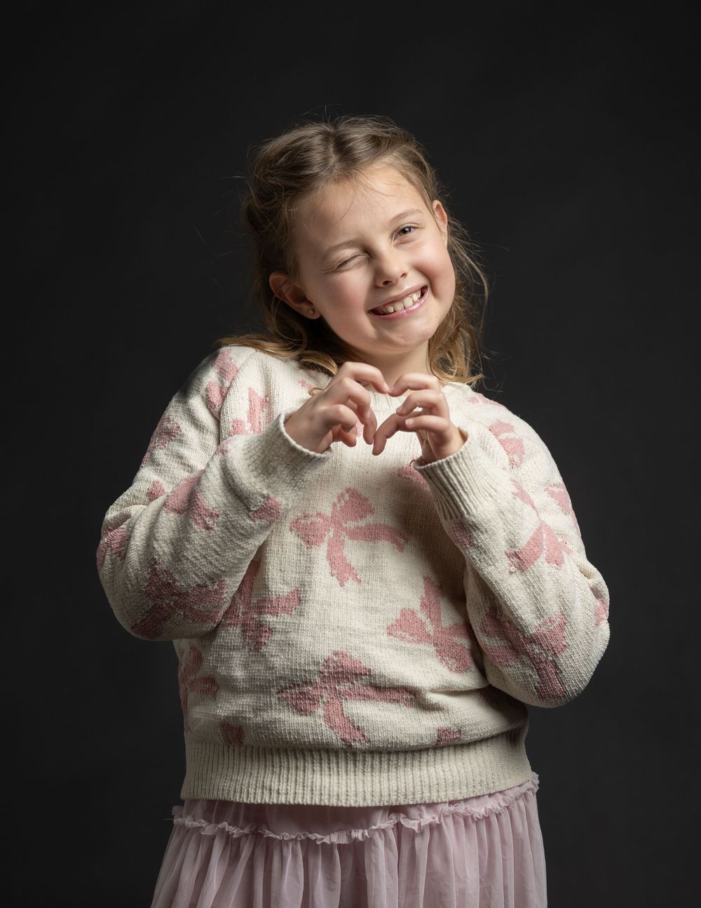 Cheerful young girl making heart shape with hands wearing pink floral sweater and tutu skirt.