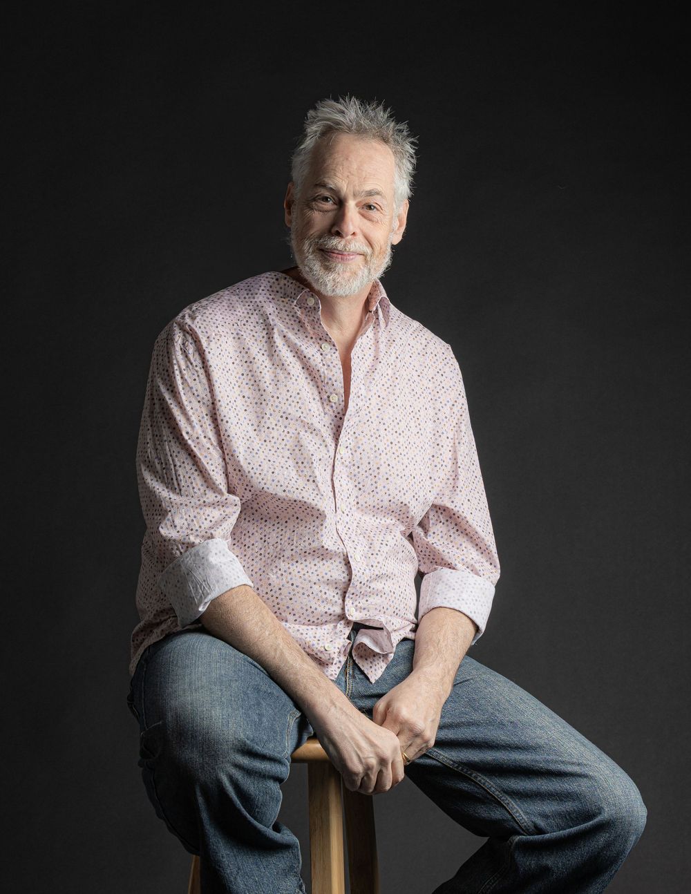 A smiling older man with gray hair and beard sits relaxed in jeans and pink shirt against dark backdrop.