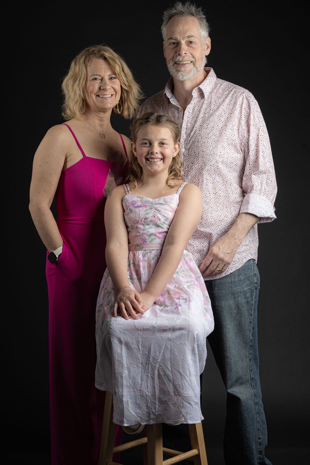 Family portrait of two adults and a young girl posing together against a dark background in a studio setting.