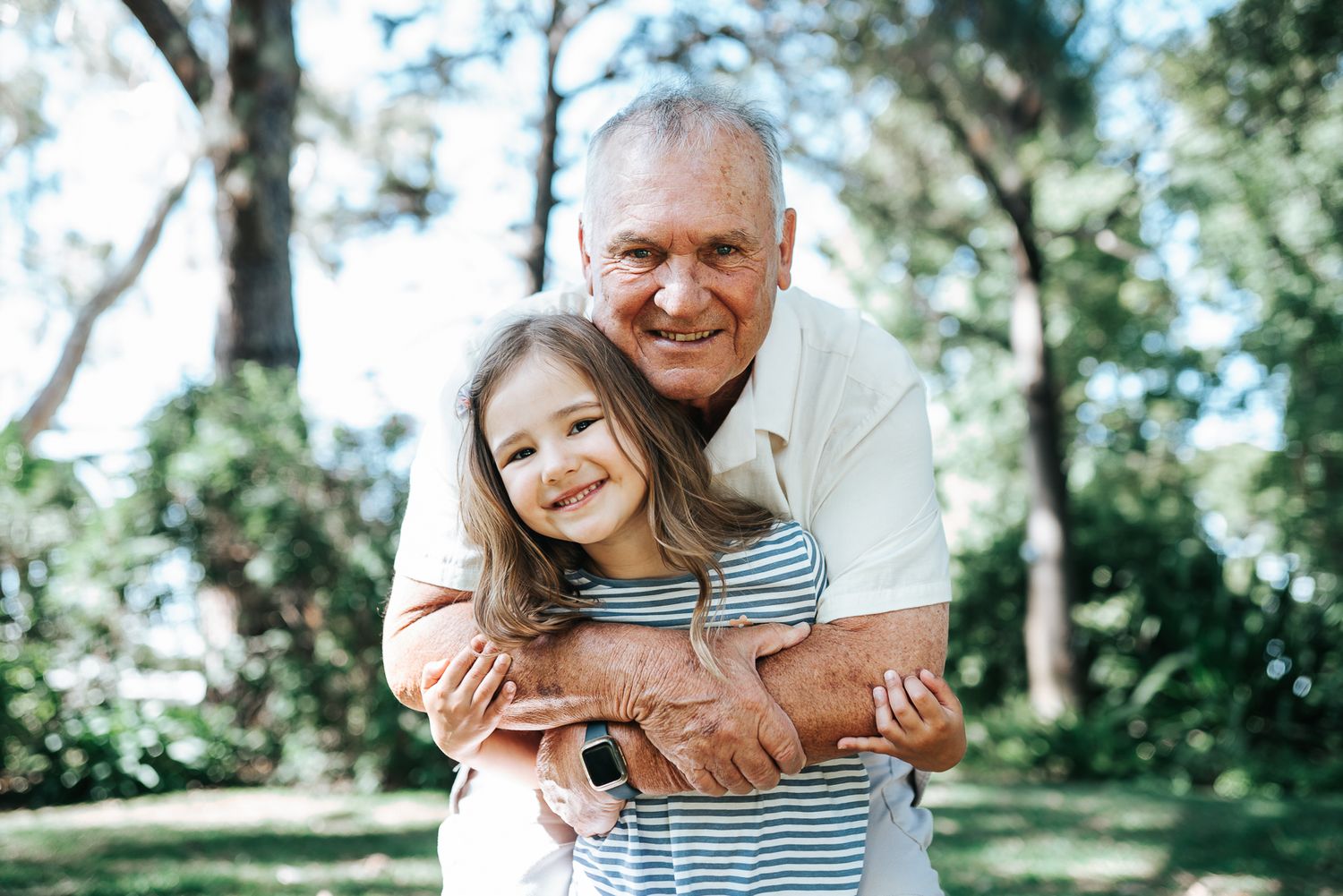 Grandfather hugging young granddaughter in a park, both smiling happily taken by Melbourne family photographer.