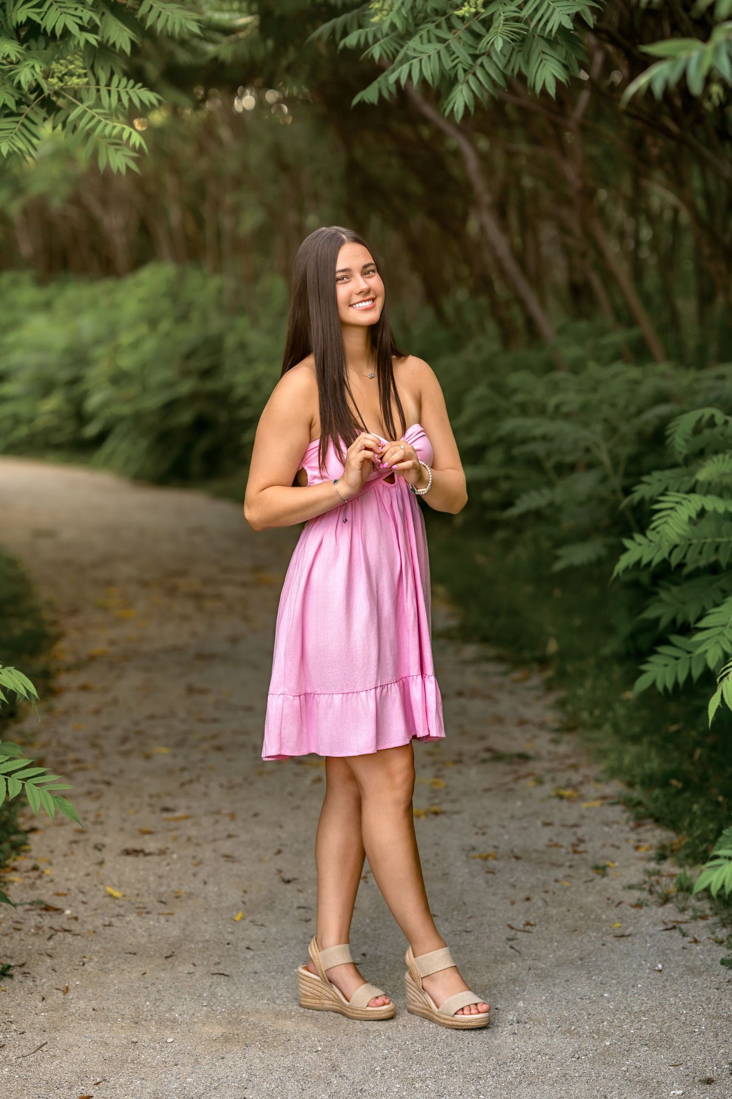 Young woman in pink sundress and wedge sandals smiling on a garden path surrounded by lush green foliage.